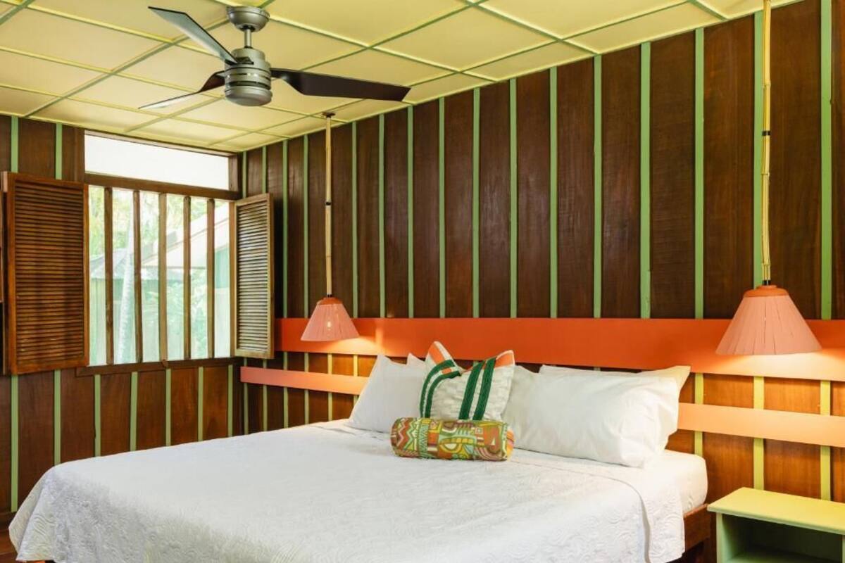 Ein Hotelzimmer mit Bett, Deckenventilator, Fenster mit Rollläden und einer Nachttischlampe.