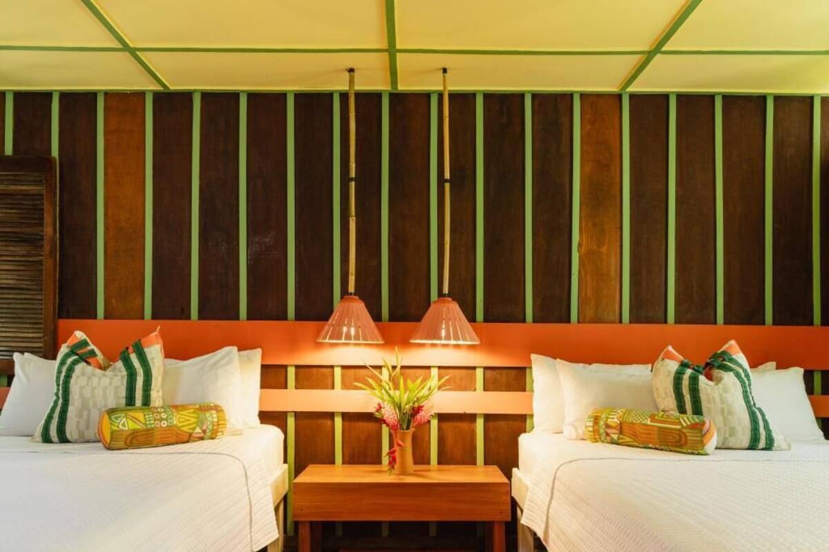 Ein Hotelzimmer mit zwei Betten, einem hölzernen Kopfteil, einem kleinen Tisch mit Blumenvase und zwei Pendelleuchten.