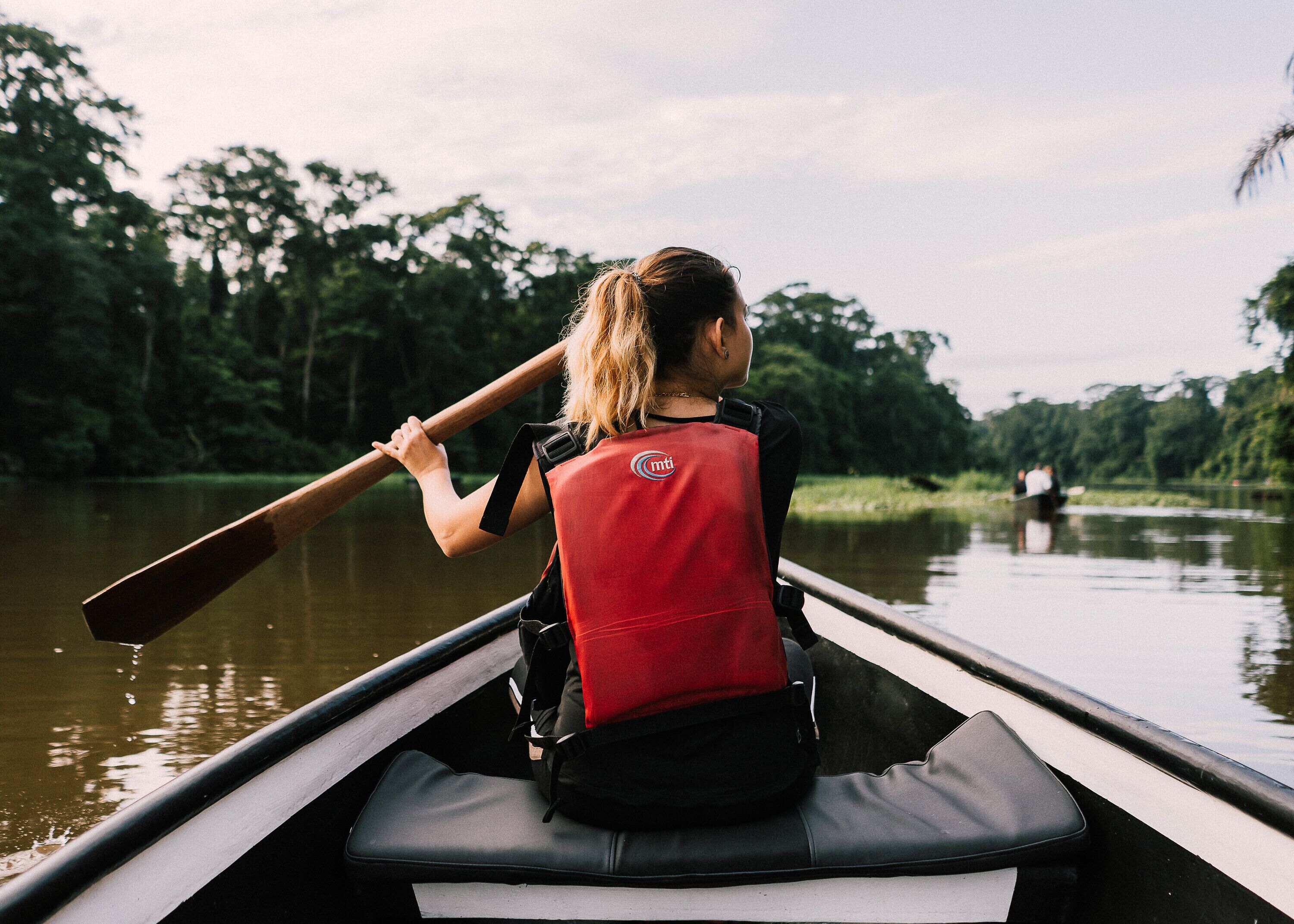 Una persona en una canoa, con chaleco salvavidas rojo, rema en un río tranquilo bordeado de árboles.