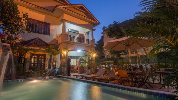 Zona de piscina con sillones de descanso y sombrillas, rodeada por un edificio con balcón.
