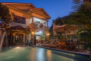 Zona de piscina con sillones de descanso y sombrillas, rodeada por un edificio con balcón.