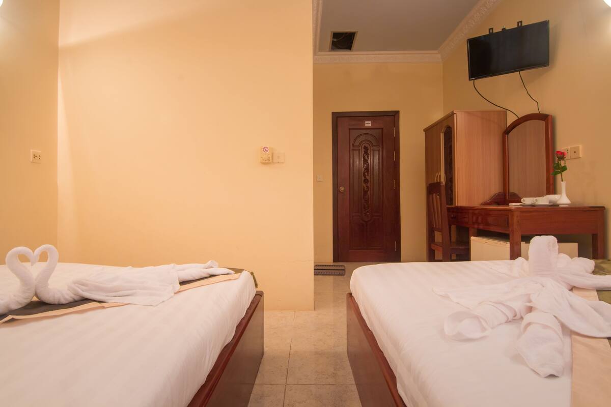 A hotel room with two beds, a wooden wardrobe, a flat-screen TV mounted on the wall, and a small desk with a vase of flowers.