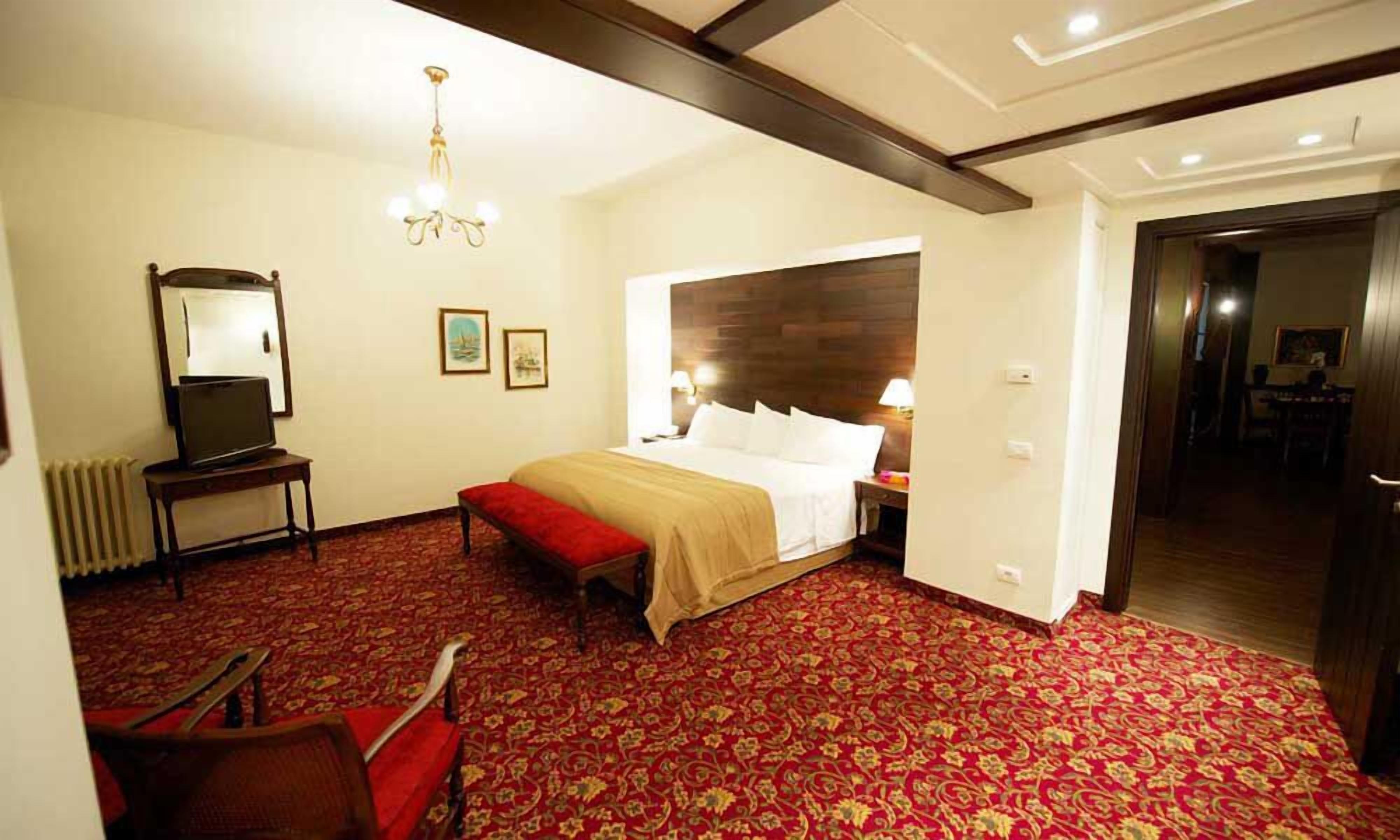 A hotel room with a large bed, a red carpet, a wooden headboard, a desk with a chair, a mirror, and a chandelier.