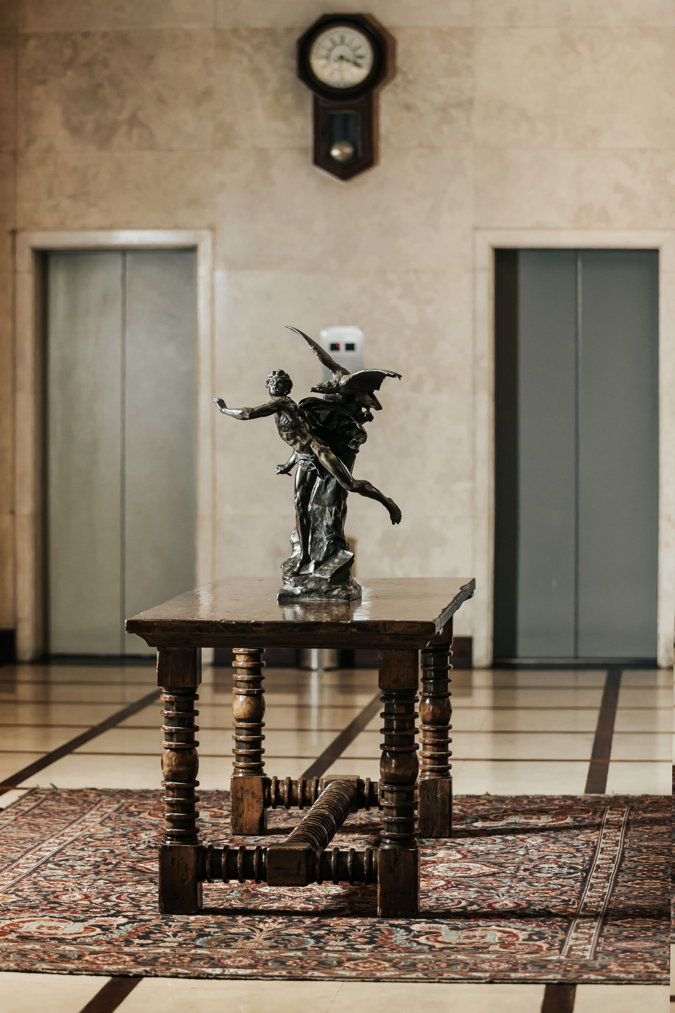 A statue on a table in a lobby with a clock on the wall and doors in the background.