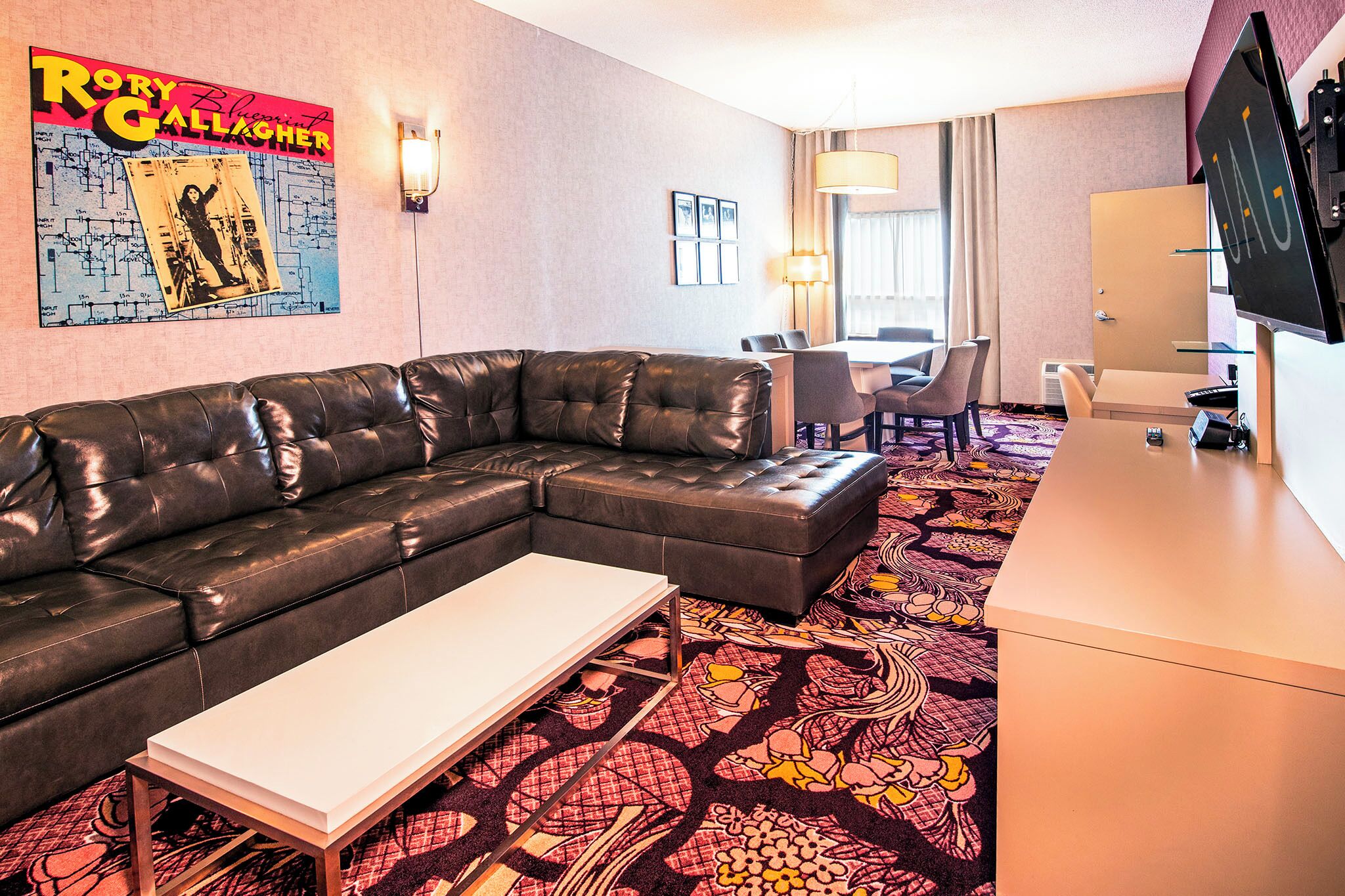 A hotel room with a sectional sofa, a small table, a patterned carpet, a dining area with chairs, and a TV on a stand.