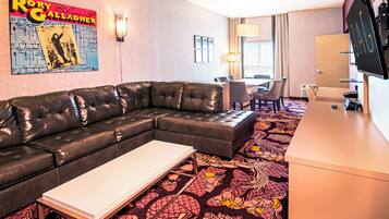 A hotel room with a sectional sofa, a small table, a patterned carpet, a dining area with chairs, and a TV on a stand.