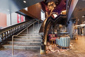 A grand staircase with a black and gold railing, a plush seating area with patterned upholstery, and a modern bar/lounge with a unique ceiling design.