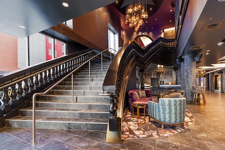 A grand staircase with a black and gold railing, a plush seating area with patterned upholstery, and a modern bar/lounge with a unique ceiling design.. Jag Boutique Hotel