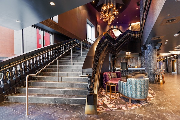 A grand staircase with a black and gold railing, a plush seating area with patterned upholstery, and a modern bar/lounge with a unique ceiling design.