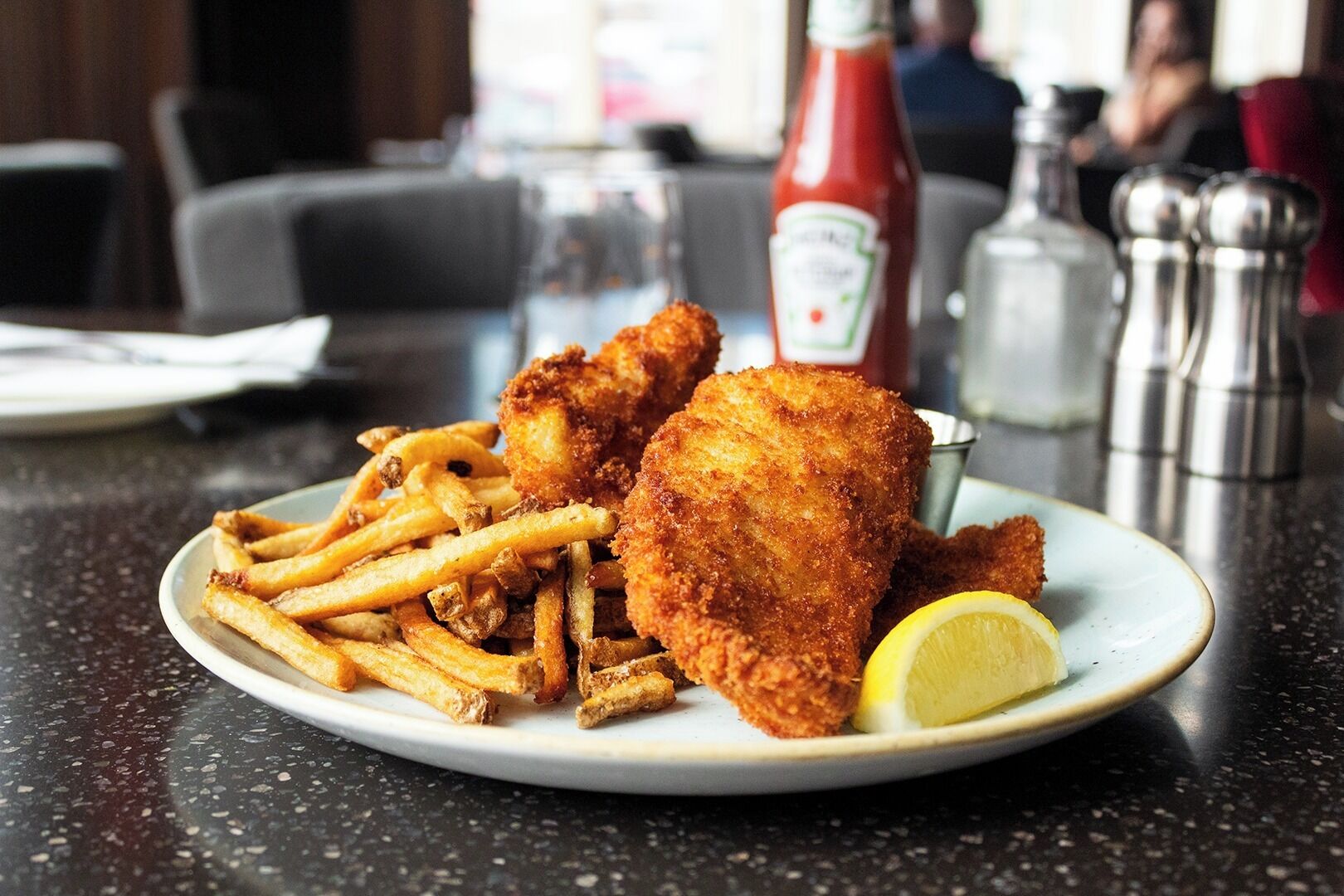 A plate of breaded fish fillets, fries, and a lemon wedge on a table with condiments and salt shakers.