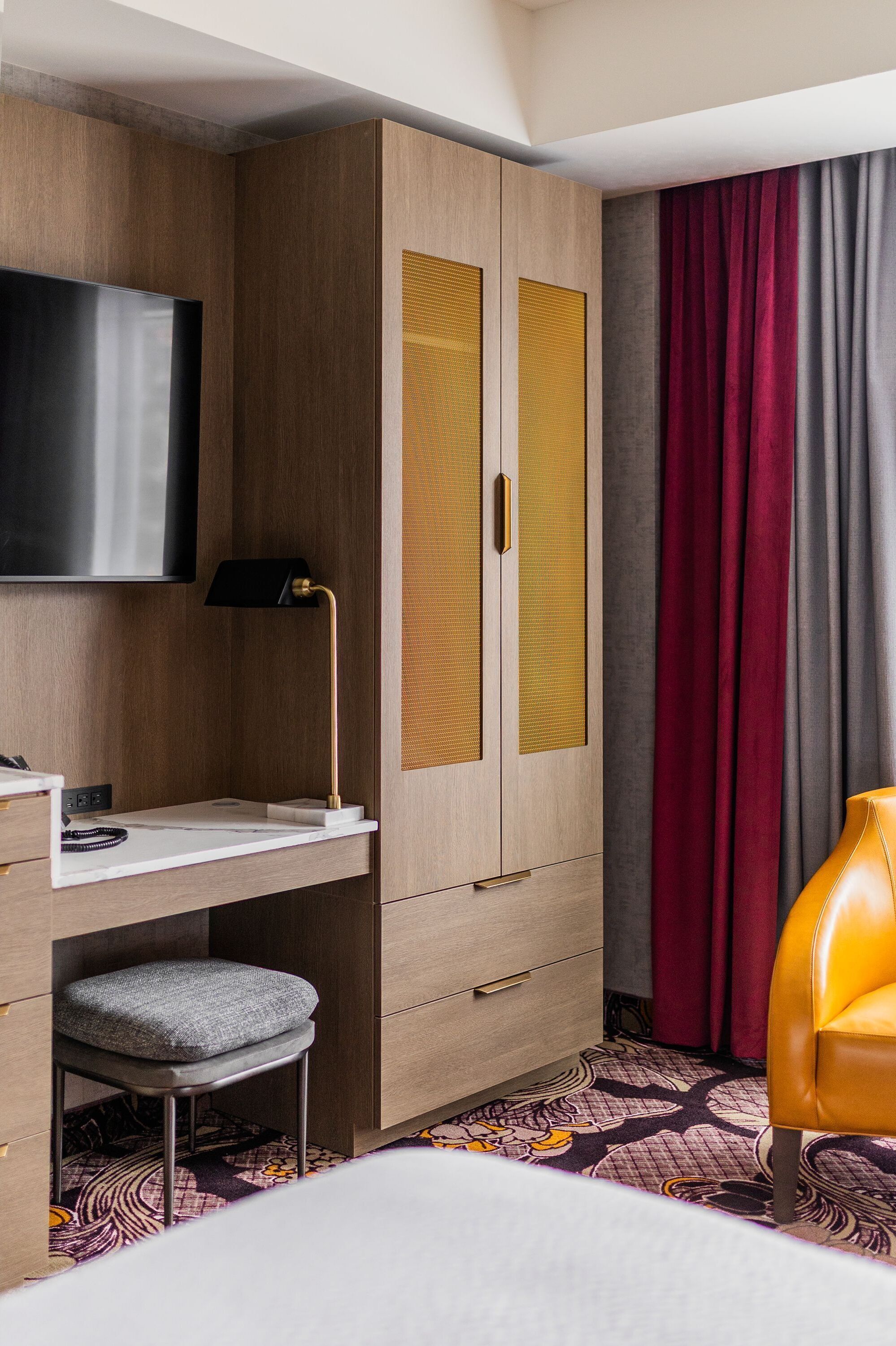 A hotel room with a wooden wardrobe, a flat-screen TV, a desk with a lamp, a cushioned stool, and a yellow armchair.
