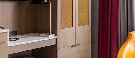 A hotel room with a wooden wardrobe, a flat-screen TV, a desk with a lamp, a cushioned stool, and a yellow armchair.