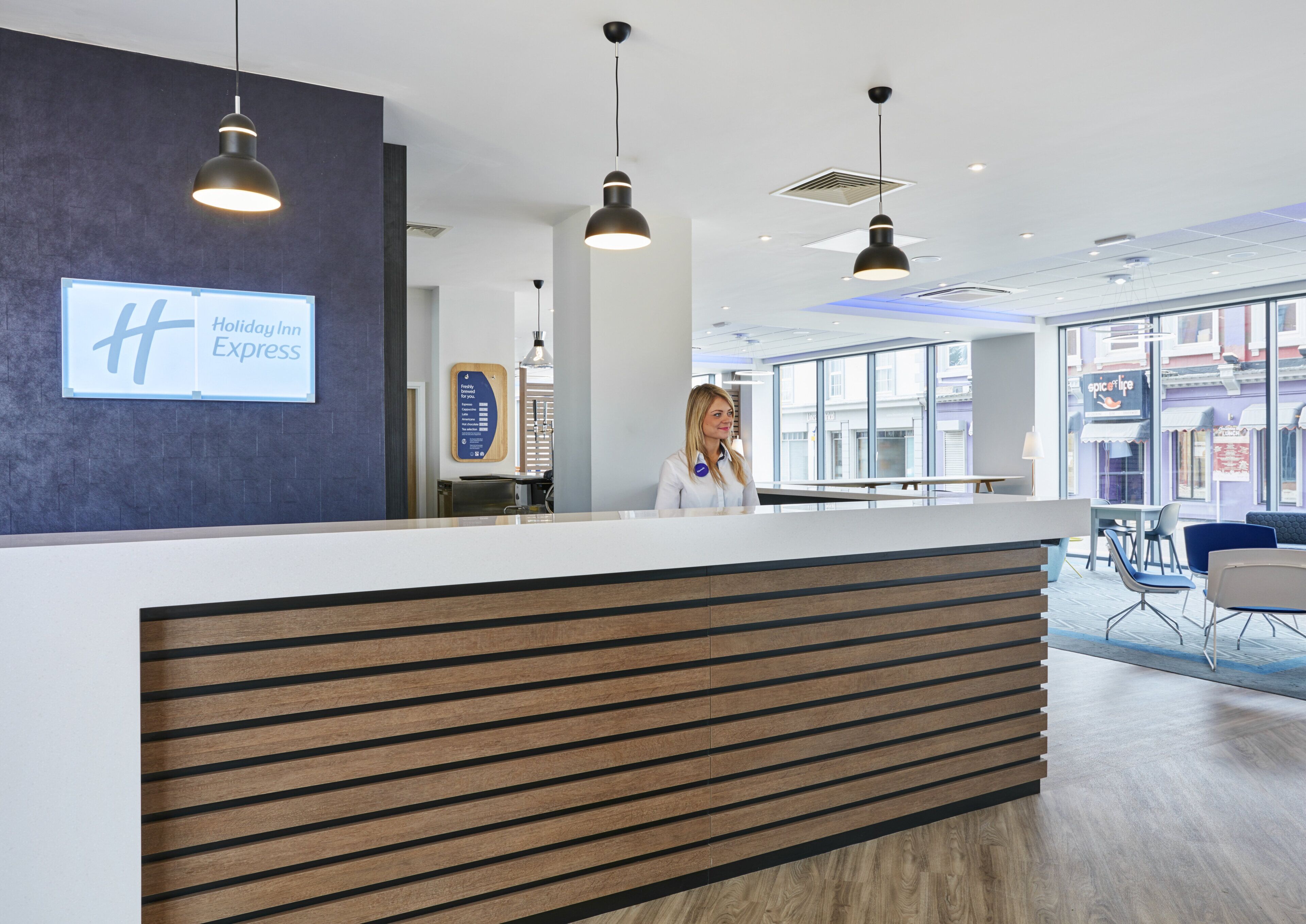 A hotel reception desk with a modern design, a sign for Holiday Inn Express, and a view of the street through large windows.