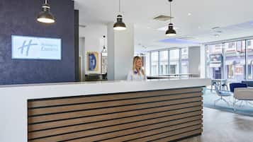A hotel reception desk with a modern design, a sign for Holiday Inn Express, and a view of the street through large windows.