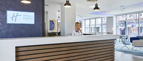 A hotel reception desk with a modern design, a sign for Holiday Inn Express, and a view of the street through large windows.
