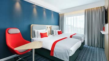 Quarto de hotel com uma cama grande, duas cadeiras vermelhas, uma mesa pequena e uma TV na parede.