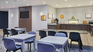 Quarto de hotel moderno com área de refeições, televisão de tela plana fixada em um painel de madeira, bancada de cozinha com cafeteira e espaço para buffet.
