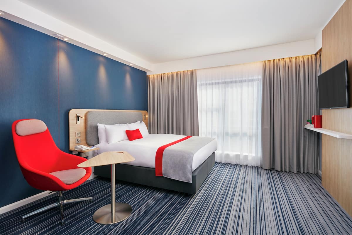 A hotel room with a bed, a red armchair, a bedside table, a TV, and a window with curtains.