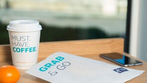 Een koffiebeker met de tekst 'MUST HAVE COFFEE', een smartphone en een papiertje met 'GRAB & GO' op een houten ondergrond.