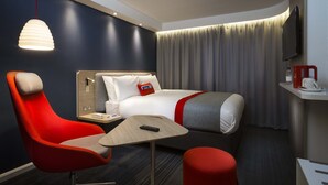 Een moderne hotelkamer met een bed, een rode fauteuil, een klein tafeltje, een rode poef, een televisie en een gordijn.