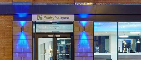 Exterior de un Holiday Inn Express con entrada de vidrio, puertas automáticas y un área de estar visible a través de las ventanas.