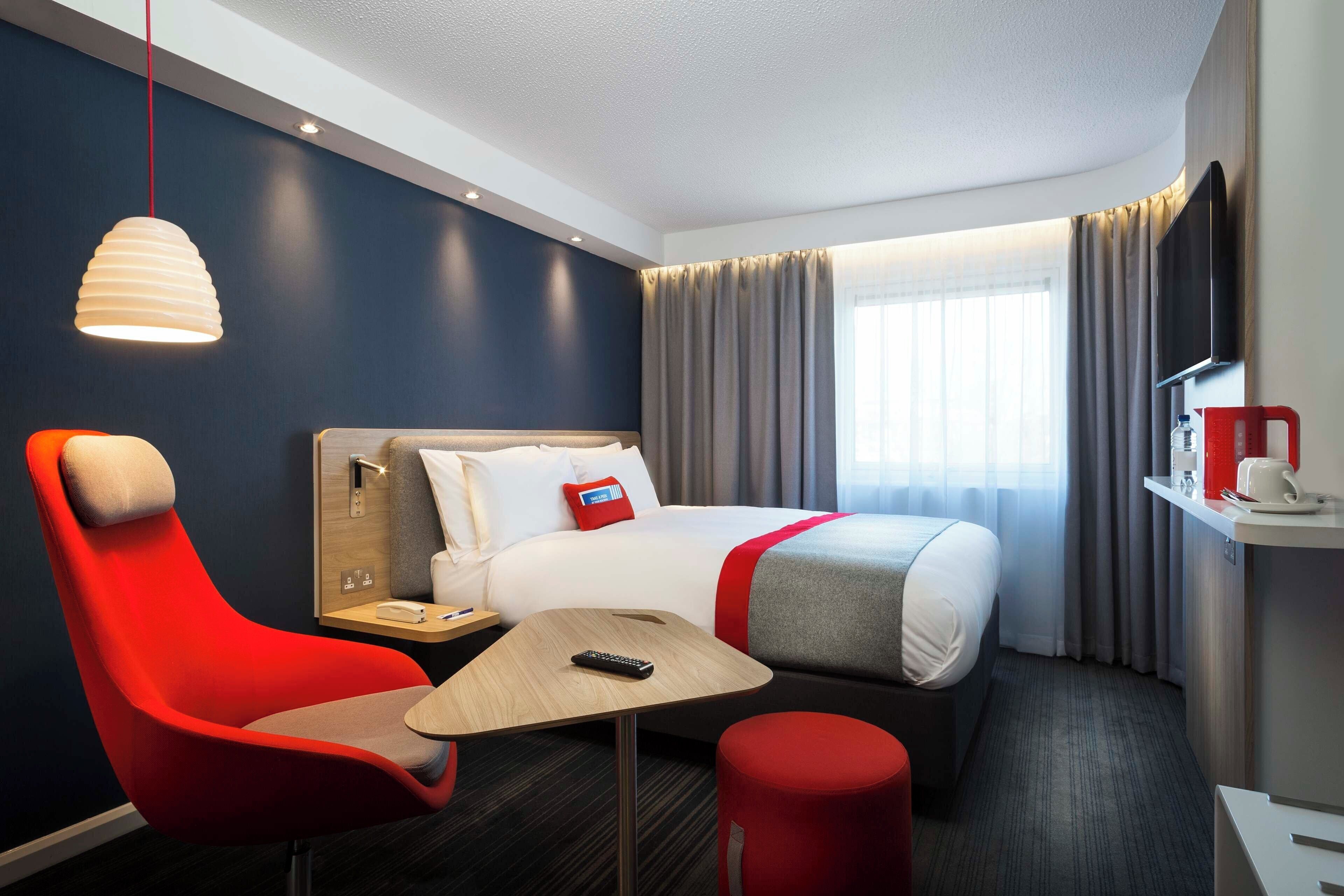 Une chambre d’hôtel avec un lit, un fauteuil rouge, une petite table, une télévision et une fenêtre avec des rideaux.