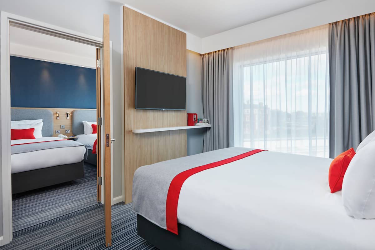 A hotel room with a large bed, a TV mounted on the wall, a small table with a red cup, and a window with grey curtains.