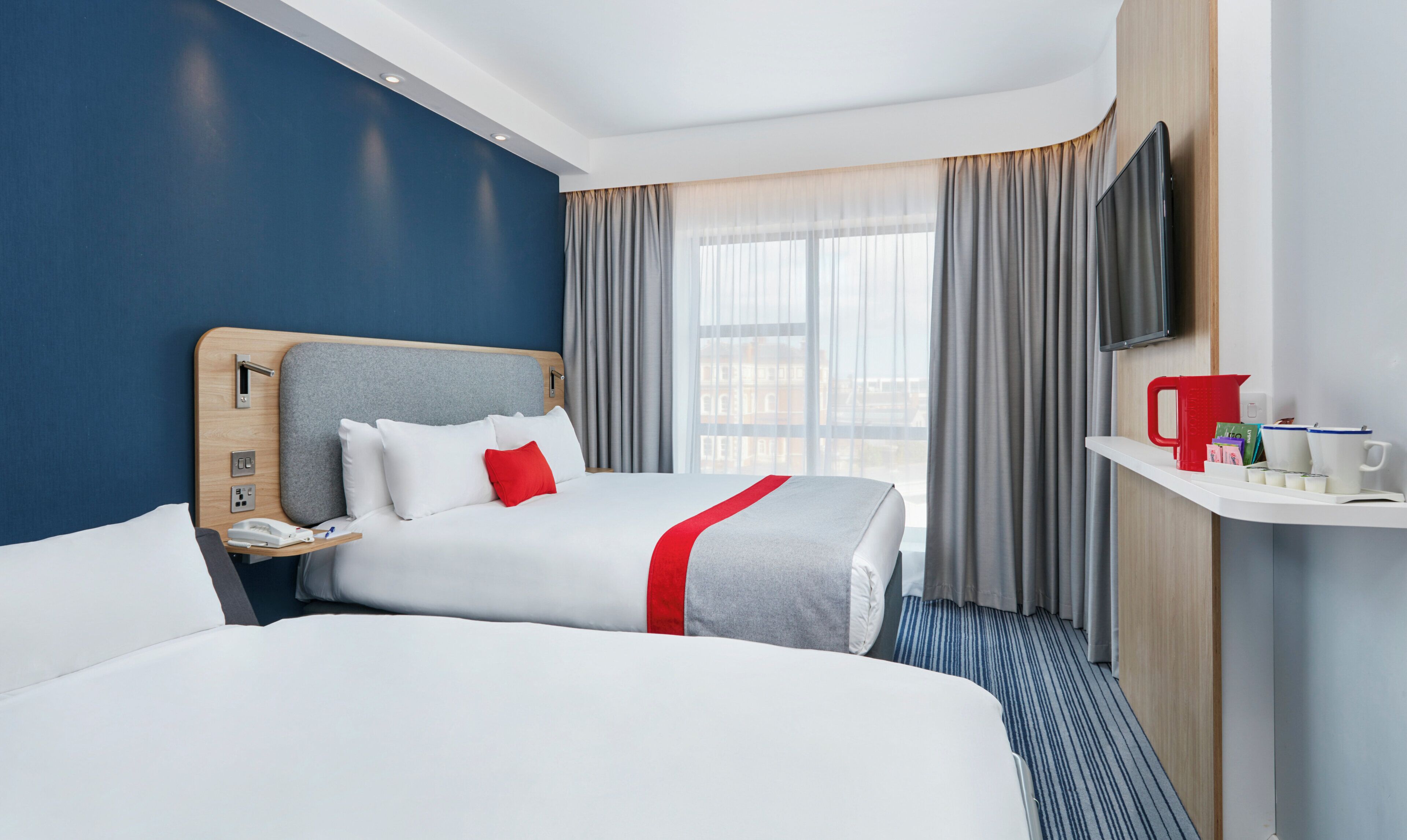 A hotel room with two beds, a TV, a bedside table with a phone, and a window with curtains.