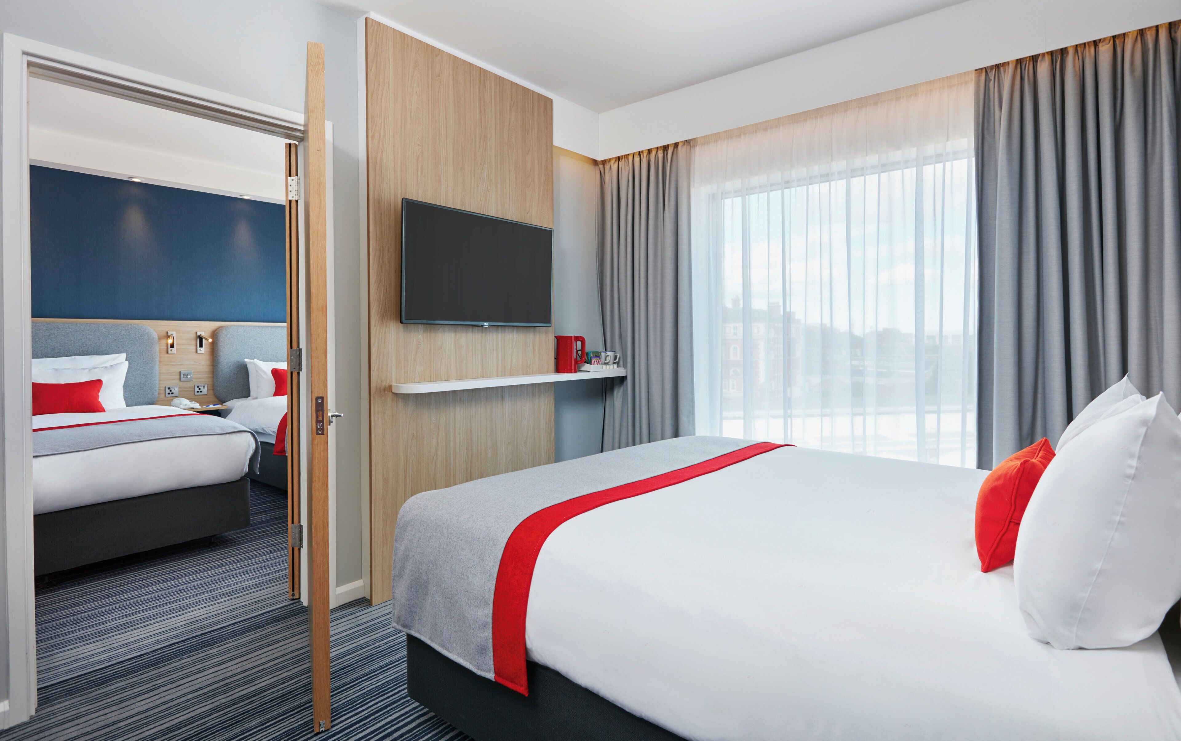 A hotel room with a bed, a television, a window with curtains, and a door leading to another room.