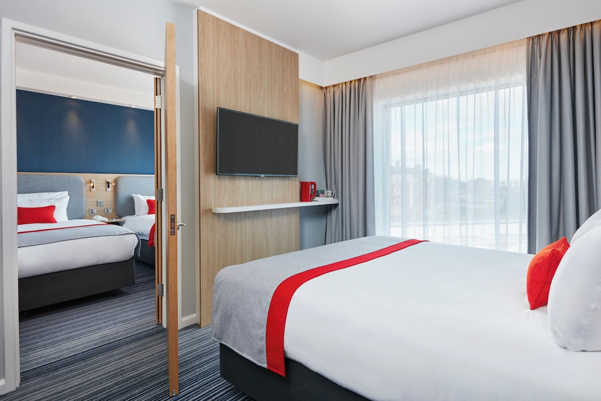A hotel room with a bed, a television, a window with curtains, and a door leading to another room.