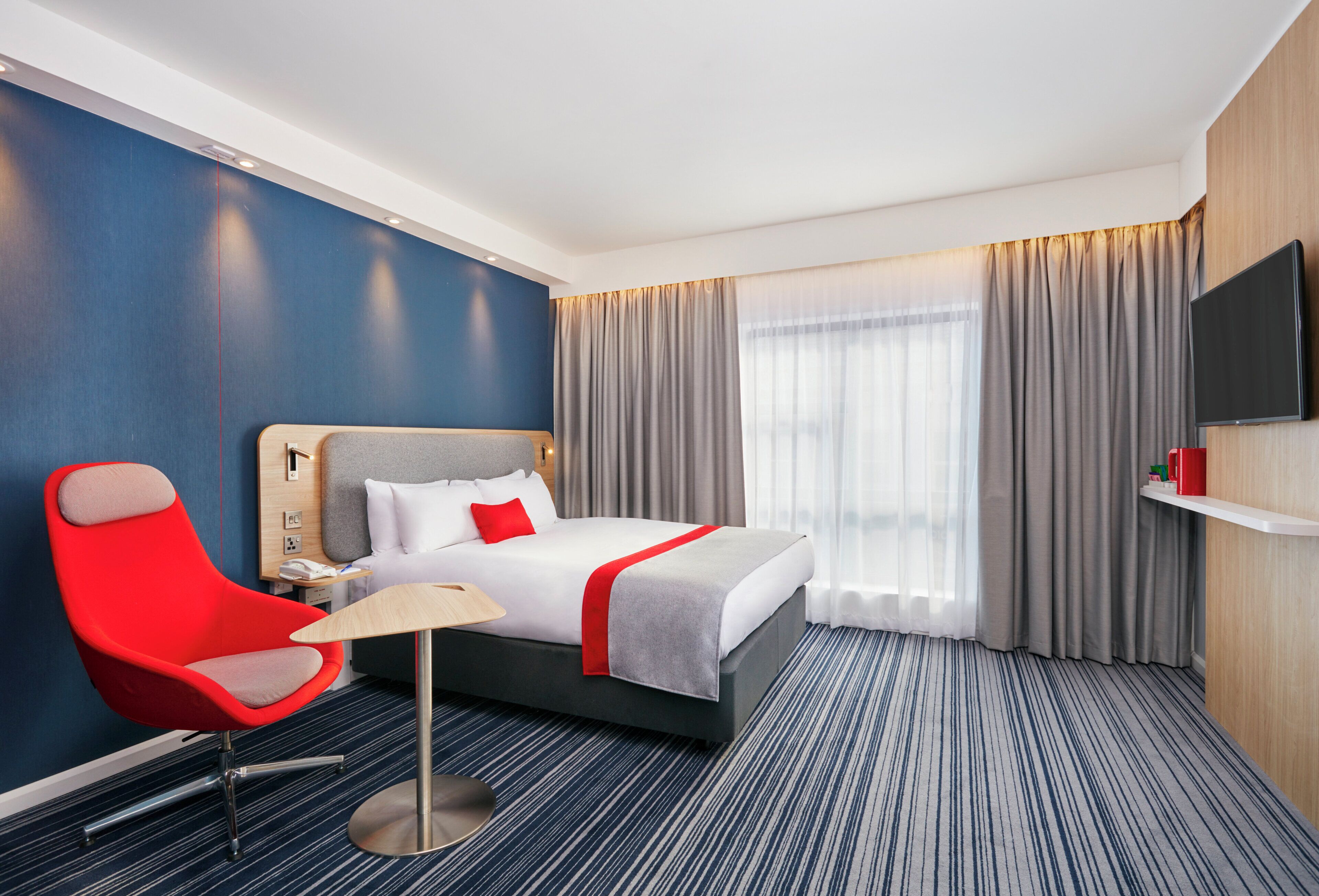 Quarto de hotel com cama, uma cadeira vermelha, escrivaninha, televisão e uma janela grande com cortinas.
