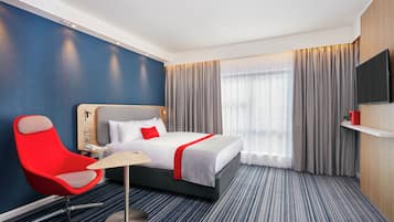 Quarto de hotel com cama, uma cadeira vermelha, escrivaninha, televisão e uma janela grande com cortinas.