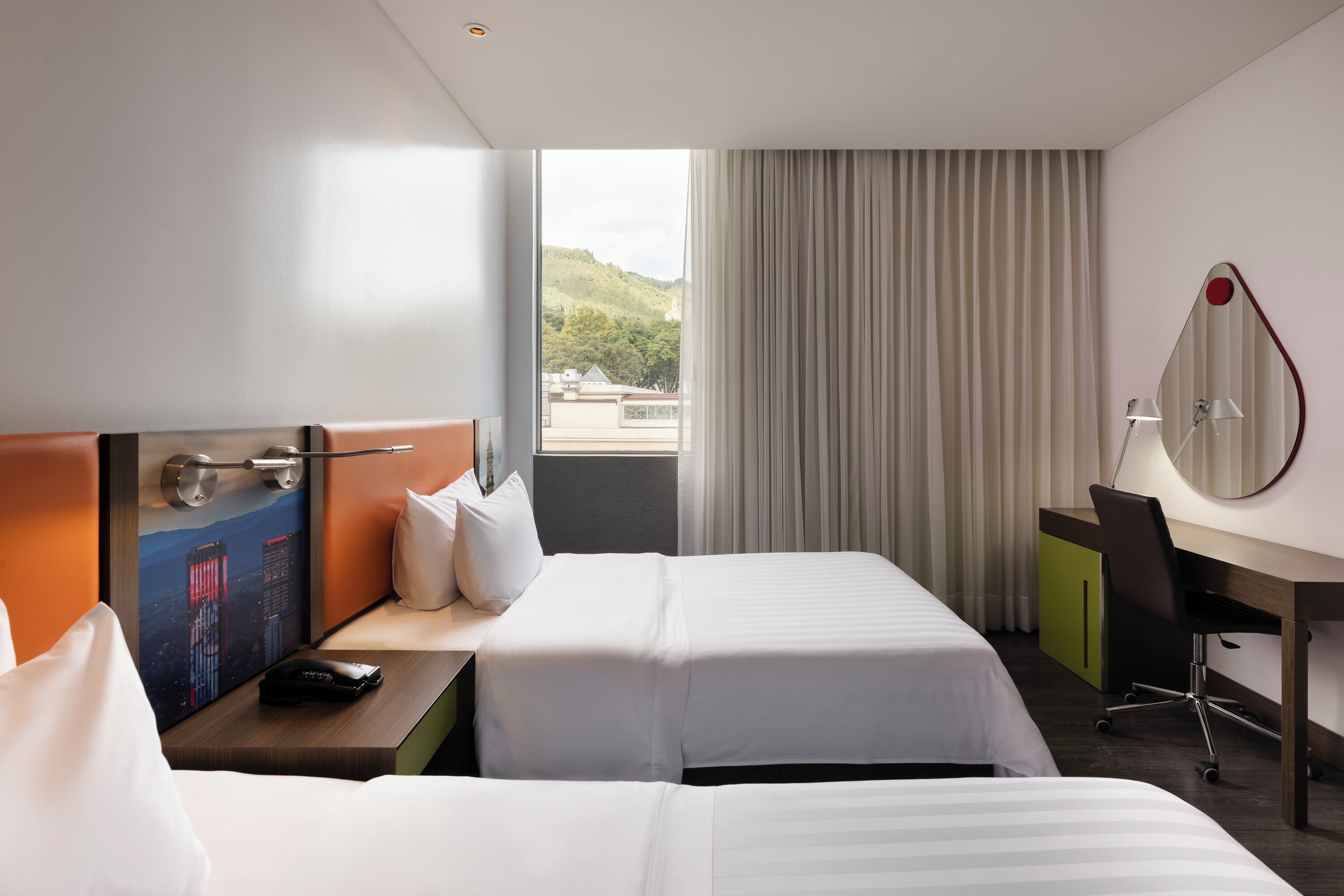 Quarto de hotel com duas camas, uma escrivaninha, uma cadeira, um abajur e vista para a cidade.
