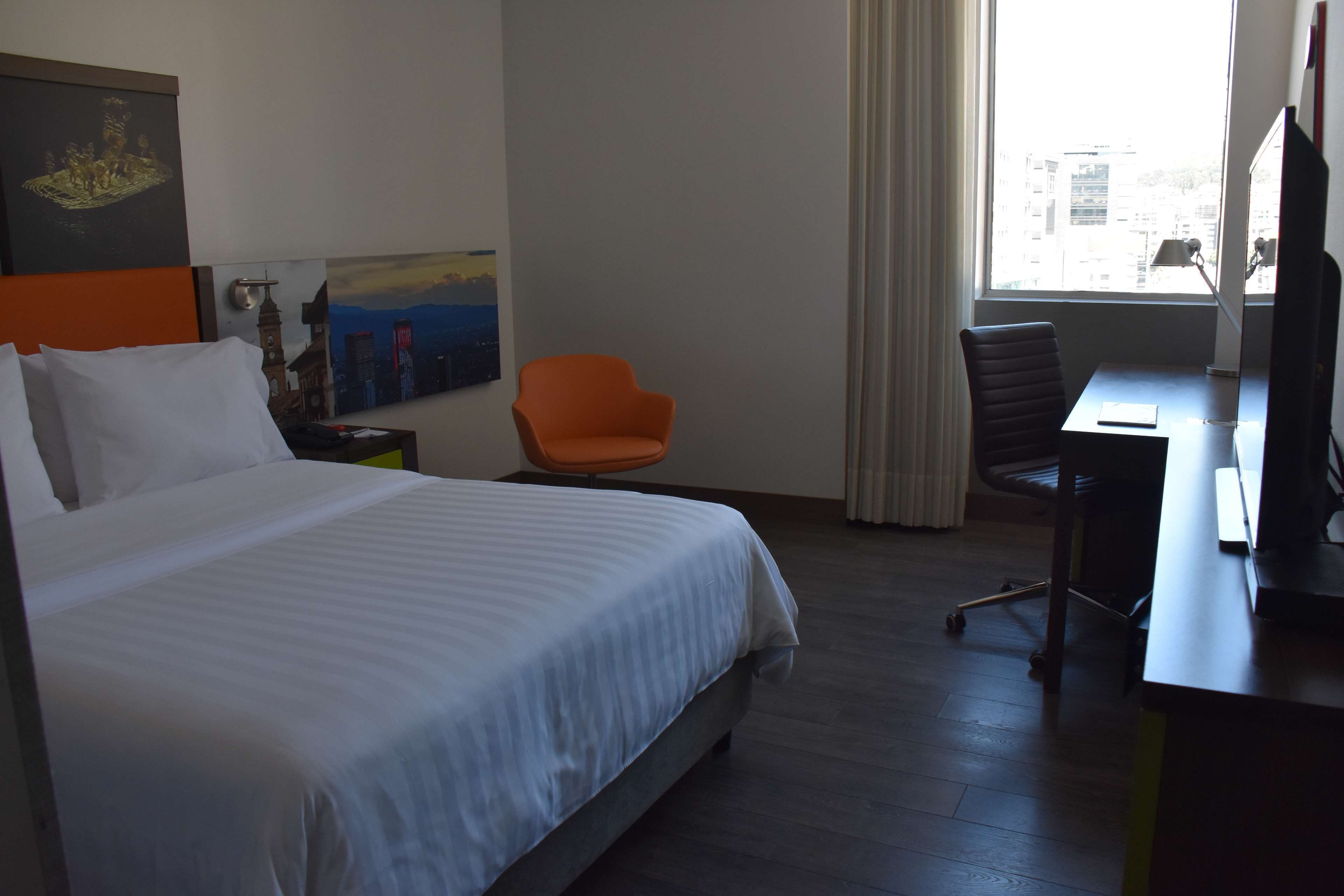 Ein Hotelzimmer mit Bett, einem orangefarbenen Sessel, einem Schreibtisch und einem großen Fenster.