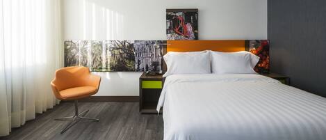 A hotel room with a bed, a chair, a nightstand, and artwork on the wall.
