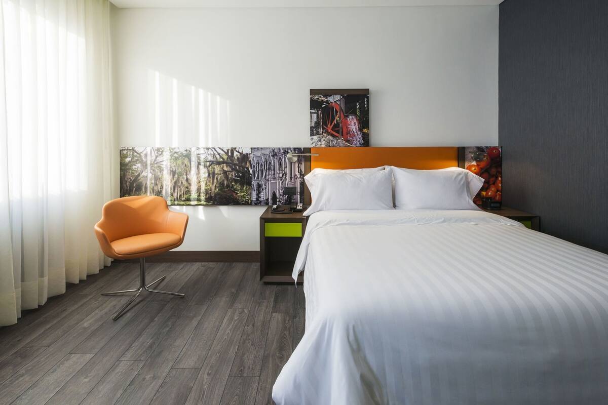 A hotel room with a bed, an orange chair, a nightstand, a painting, and a window with curtains.