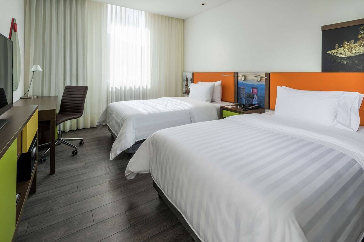 A hotel room with two beds, a desk, a chair, a lamp, and a painting on the wall.