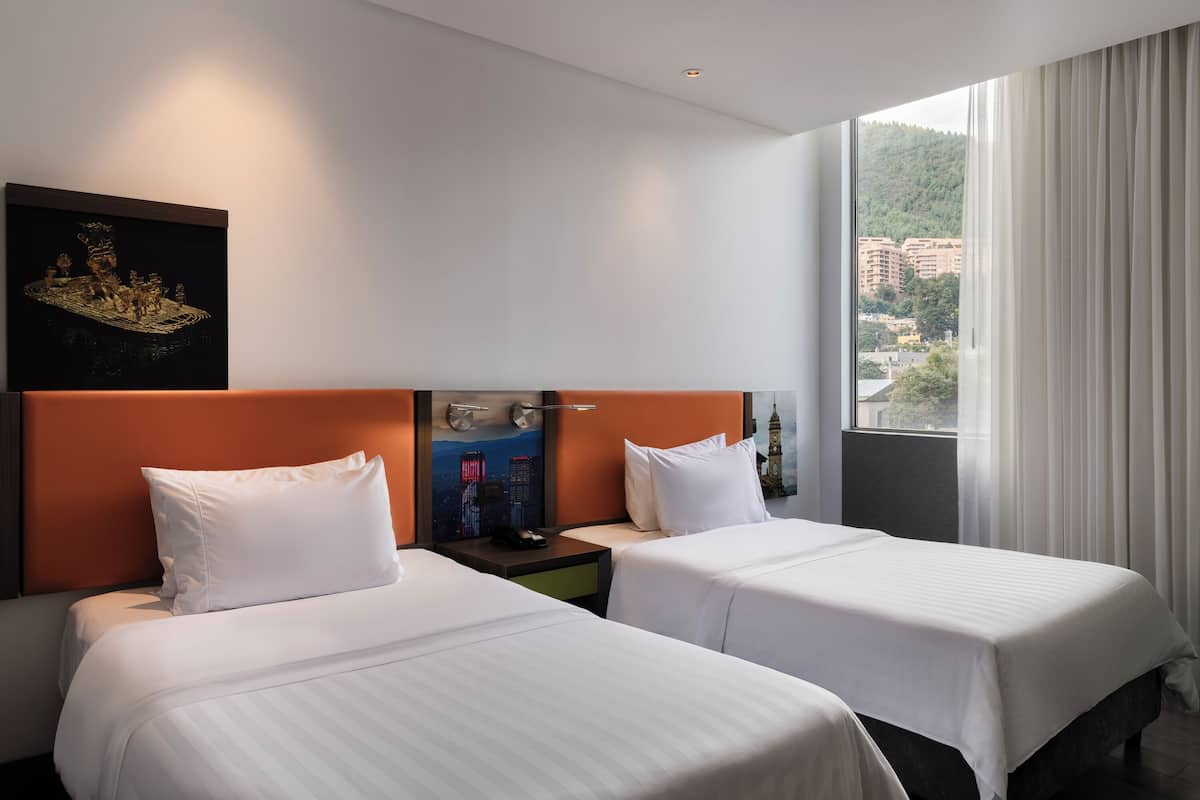 A hotel room with two beds, a nightstand, a wall-mounted clock, and a window with a view of buildings.