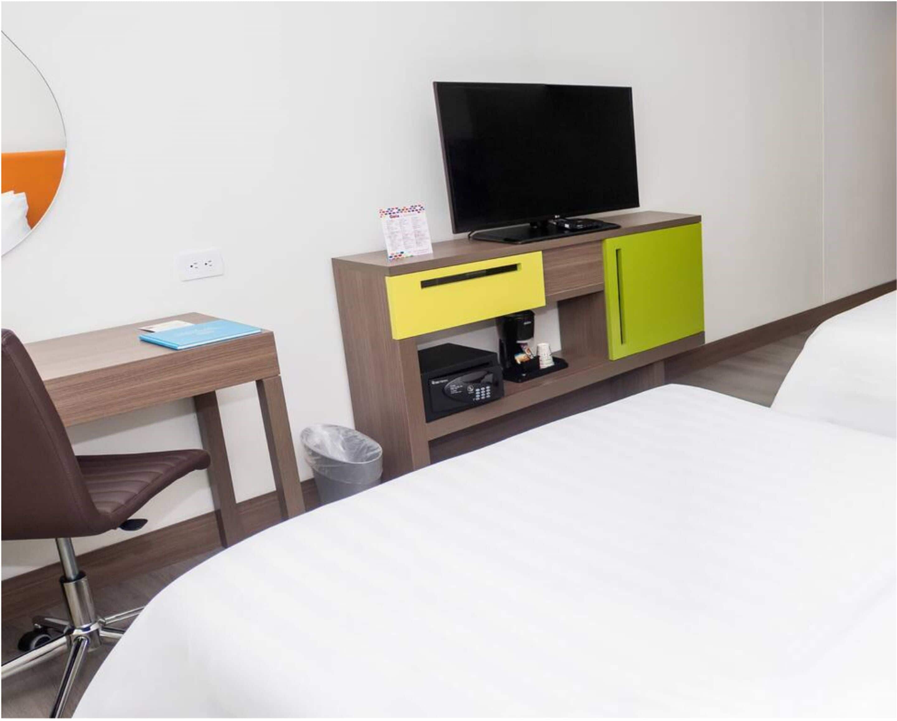A hotel room with a bed, a desk with a chair, a television, a coffee machine, and a trash bin.