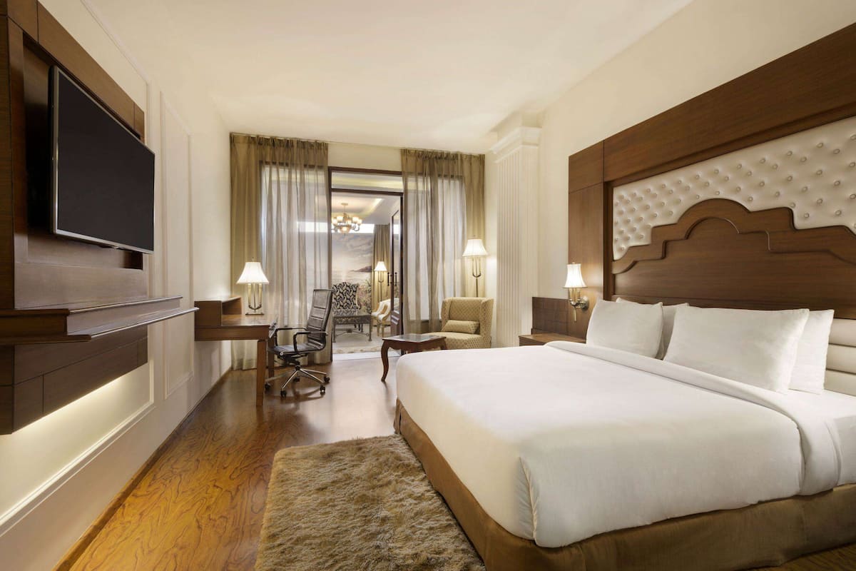 A hotel room with a large bed, a television mounted on the wall, a desk with a chair, and a view of the outside through a door.