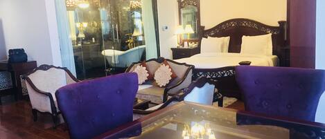 A hotel room with a bed, a glass-enclosed bathroom, a dining table with chairs, and a chandelier.