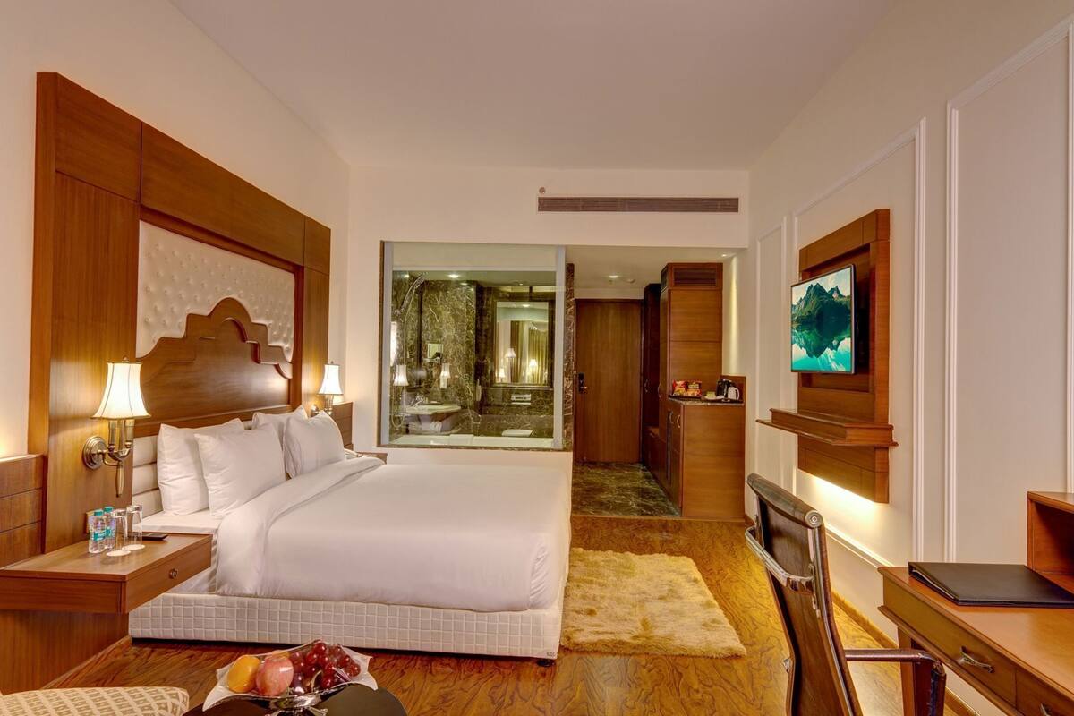 A hotel room with a large bed, a TV, a desk, a bathroom with a mirror, and a view of the bathroom through a glass door.