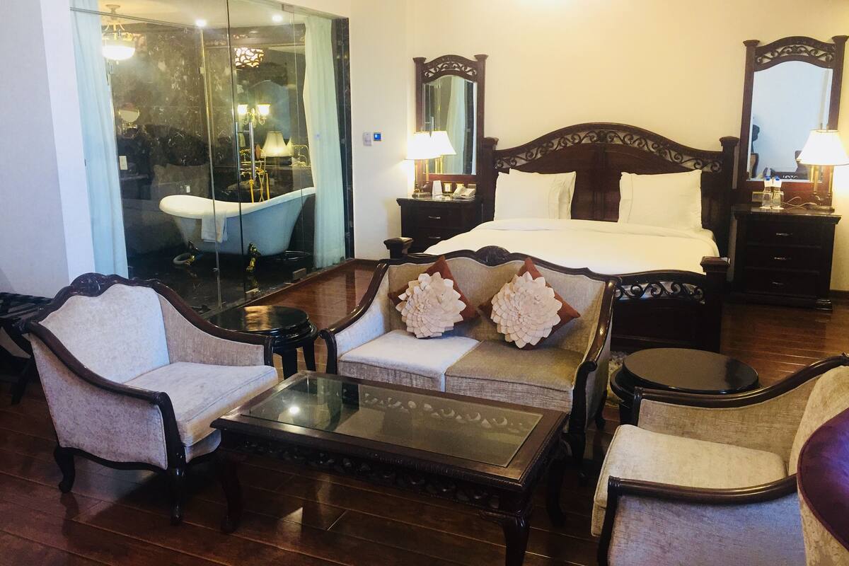 A hotel room with a large bed, two armchairs, a sofa, a glass-enclosed bathroom, and a wooden floor.