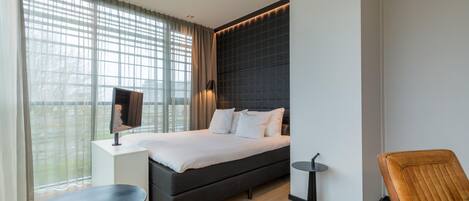 Una habitación de hotel moderna con una cama grande, una mesita, una silla, un televisor y un ventanal con cortinas.