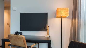 A hotel room with a desk, a chair, a flat-screen TV, a lamp, and a telephone.