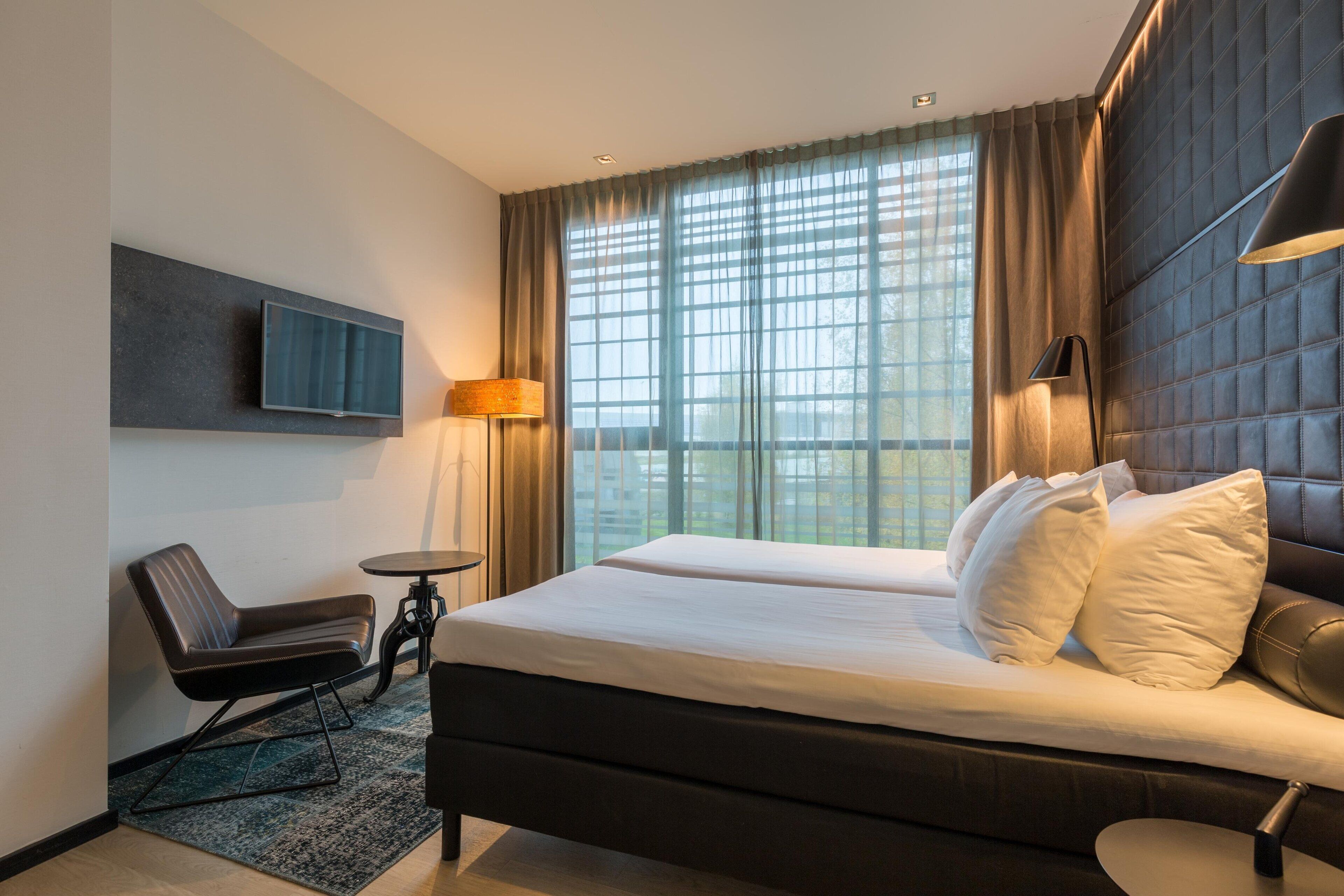 A modern hotel room with a large bed, a chair, a small table, a TV mounted on the wall, and a large window with curtains.