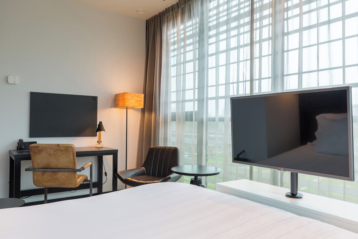 Habitación de hotel con cama, escritorio con lámpara, silla, televisor y ventanas amplias.
