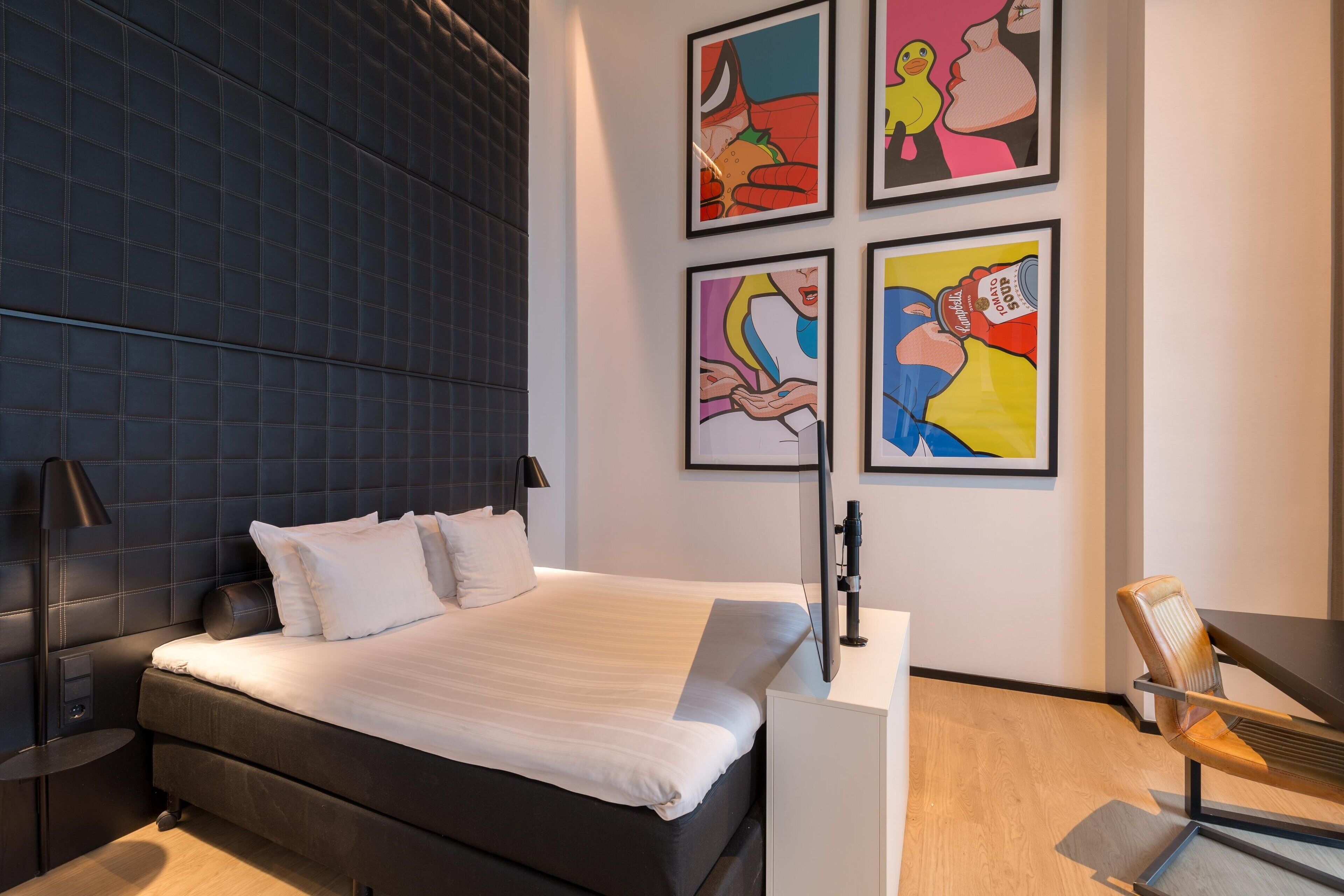 A modern hotel room with a large bed, a desk, and a chair. There are four framed artworks on the wall.