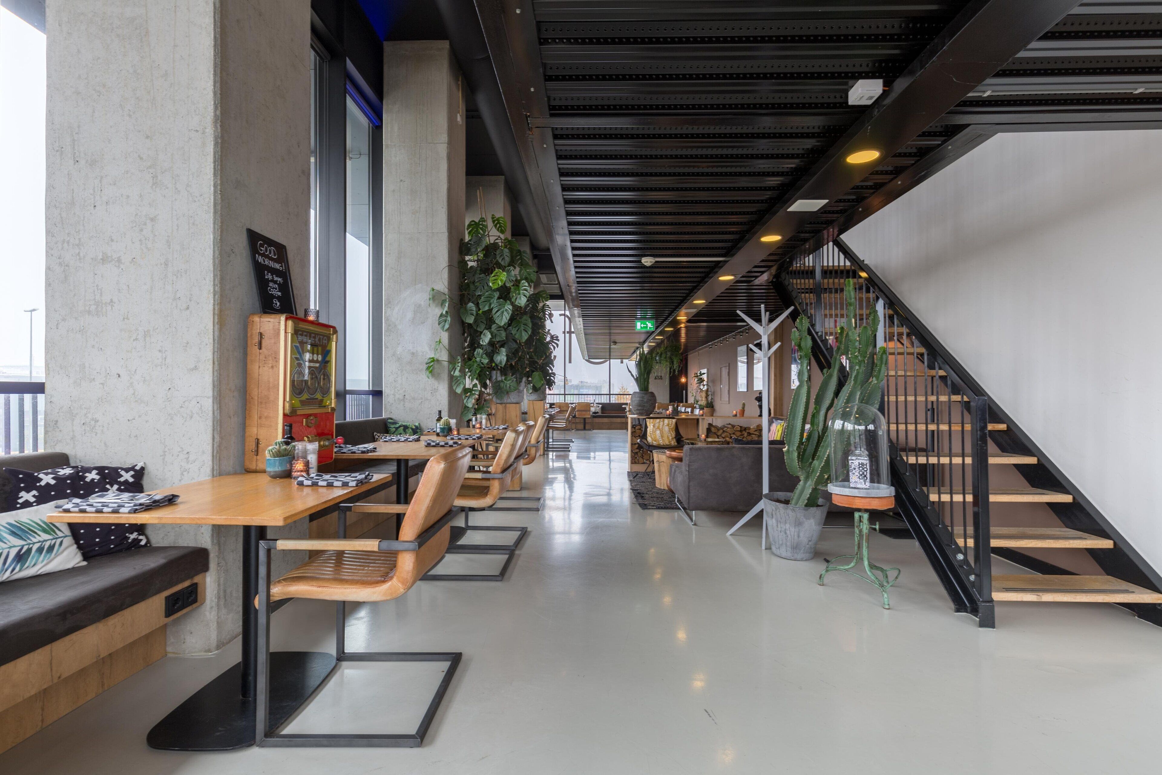 Een modern interieur met een lange eettafel, houten tafels en stoelen. Aan de rechterkant bevindt zich een trap en in het midden staat een grote plant.