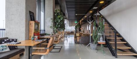 Un intérieur moderne avec un grand espace de salle à manger, des tables et des chaises en bois. Un escalier se trouve sur la droite et une grande plante est placée au centre.
